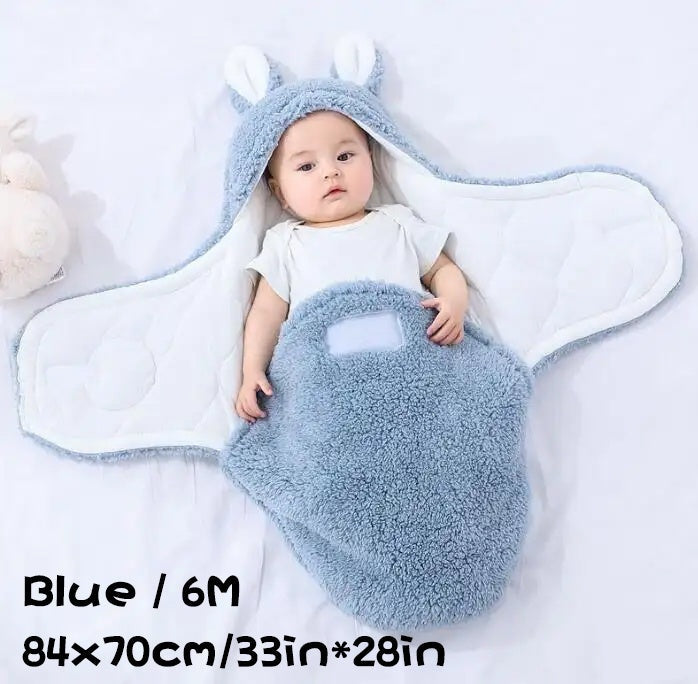 A baby wrapped in a blue fluffy swaddle sleeping bag with ear hood, labeled for size 6M and dimensions 84×70cm (33in×28in), lying on a white blanket.