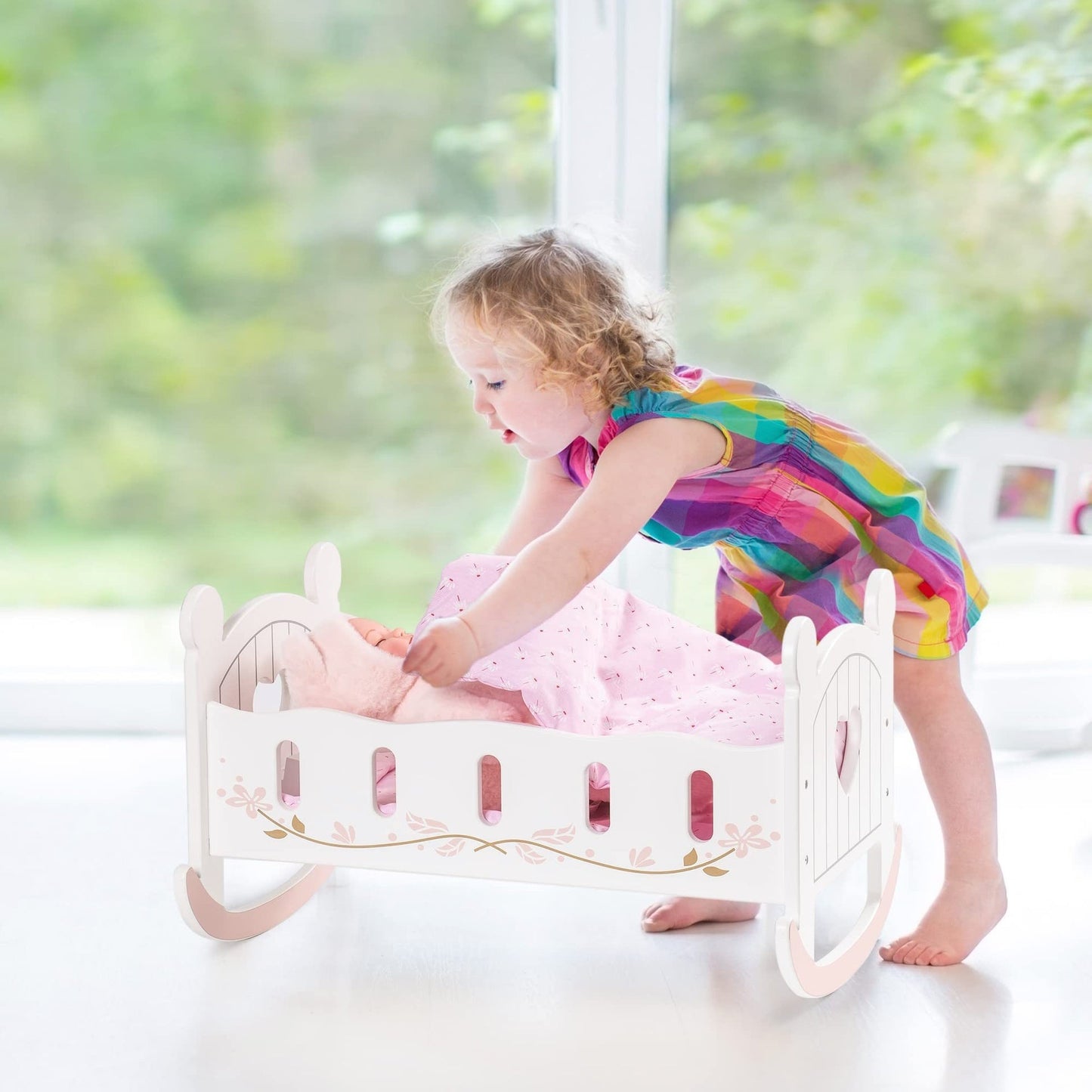 Toddler girl putting a doll to bed in a white wooden rocking cradle with heart cutouts and pink bedding