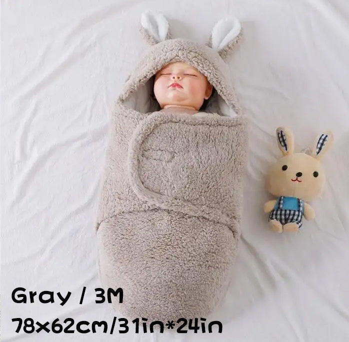 A baby wrapped in a gray fluffy swaddle sleeping bag with bunny ears, lying on a white blanket next to a plush bunny toy, with size dimensions shown.