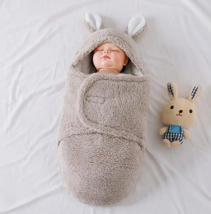 A sleeping baby wrapped in a soft, grey, fluffy swaddle with bunny ears, lying next to a plush toy on a white blanket.