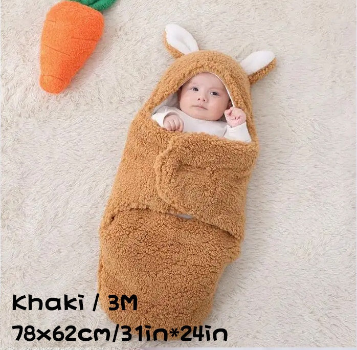 Baby wrapped in a khaki-colored fluffy swaddle sleeping bag with bunny ears, lying on a soft carpet