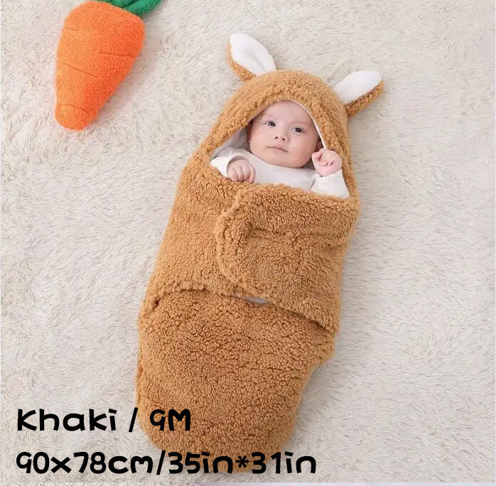Khaki baby swaddle sleeping bag in size 9M (90×78cm / 35in×31in) with baby wrapped inside, lying on a soft carpet next to a plush carrot toy.