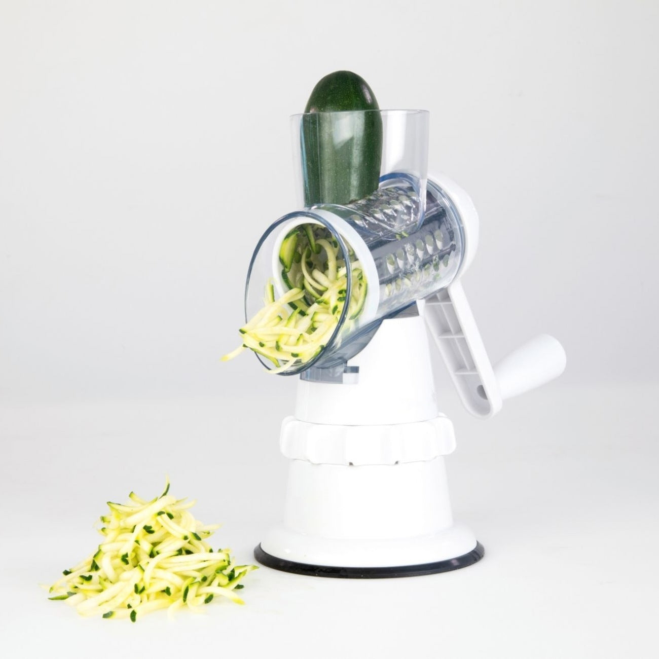 Manual vegetable shredder slicing zucchini into thin strips with rotary blade and white base.