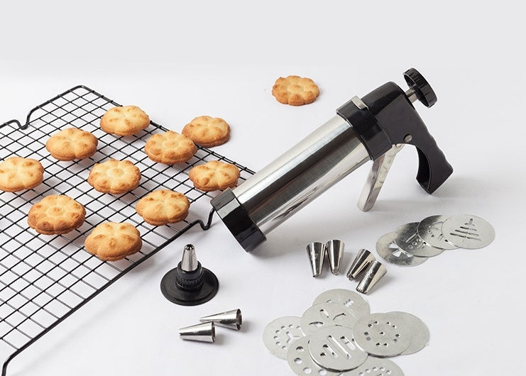 Stainless steel cookie press with icing tips and cookie discs next to baked cookies on a cooling rack