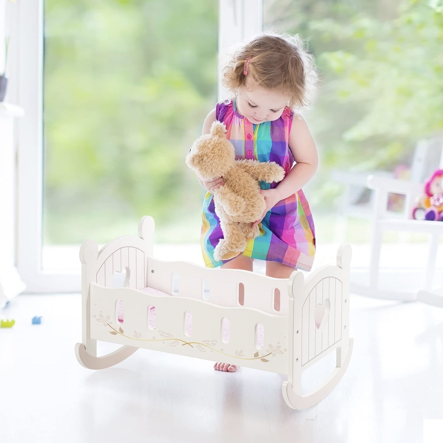 Toddler girl holding a teddy bear next to a white wooden rocking doll cradle with pink bedding