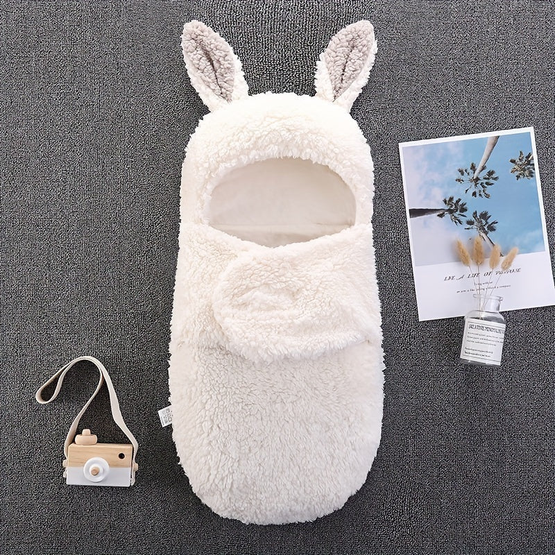 A white fluffy baby swaddle sleeping bag with bunny ears, displayed on a gray surface next to a toy camera and a decorative postcard.