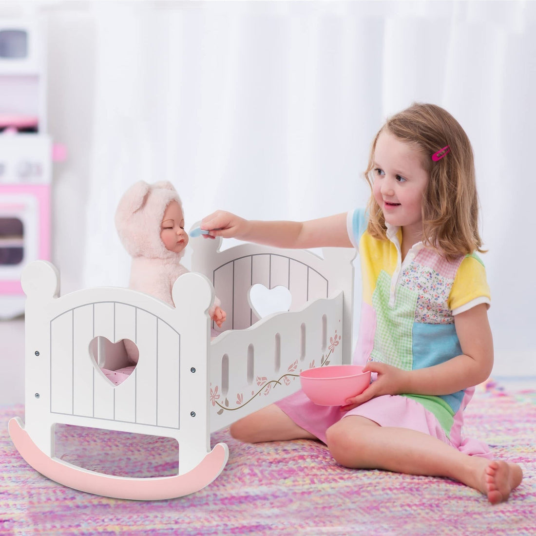 Little girl playing with a baby doll in a white wooden rocking cradle with heart-shaped cutouts and floral details.