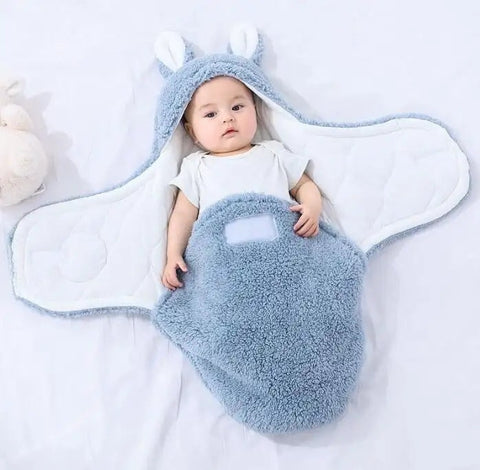 Baby lying in a soft blue fluffy swaddle sleeping bag with open wings and bunny ears hood on a white surface.