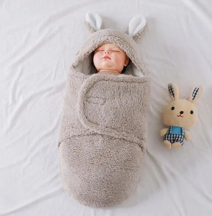 A sleeping baby wrapped in a soft, grey, fluffy swaddle with bunny ears, lying next to a plush toy on a white blanket.