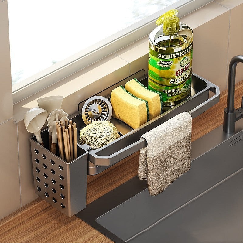 Top angle view of kitchen sink organizer with sponges, dish soap, towel, and heart-shaped utensil holder next to a black faucet