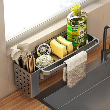 Top angle view of kitchen sink organizer with sponges, dish soap, towel, and heart-shaped utensil holder next to a black faucet