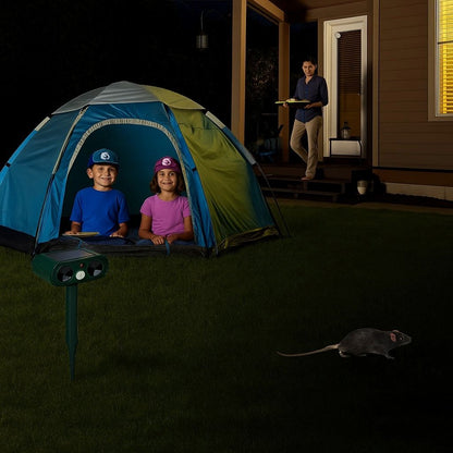 Children sitting in a tent at night near a solar ultrasonic animal repeller, with a rat approaching on the lawn and an adult bringing food from a house in the background.