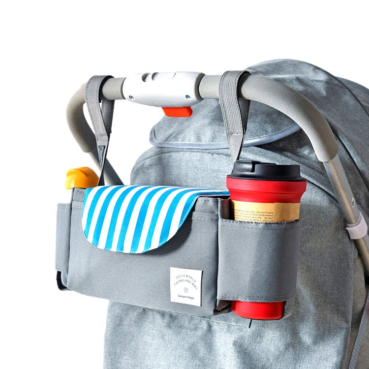 Gray stroller organizer attached to a stroller handle, featuring a blue and white striped flap and holding a travel mug and baby items, photographed on a white background.