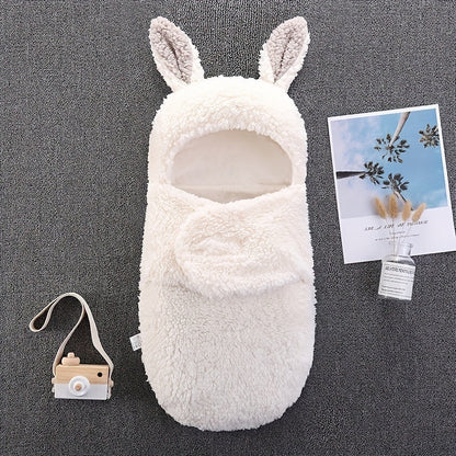 A white fluffy baby swaddle sleeping bag with bunny ears, displayed on a gray surface next to a toy camera and a decorative postcard.